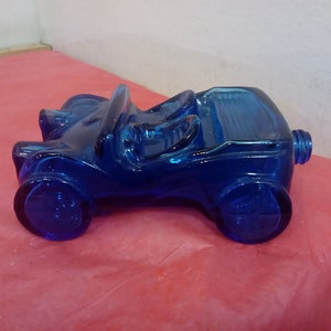 Puede incluir: Una botella de vidrio azul cobalto con forma de coche. La botella es un coche de estilo vintage con un cuerpo redondeado y una sola abertura en la parte superior.