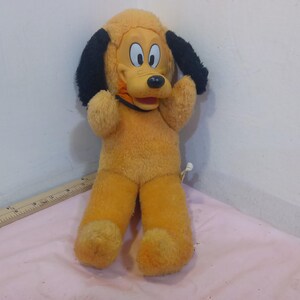 May include: A vintage plush toy of a cartoon dog with yellow fur, black ears, and a black nose. The dog has a red collar and is in a standing position with its arms raised. The toy is in good condition.
