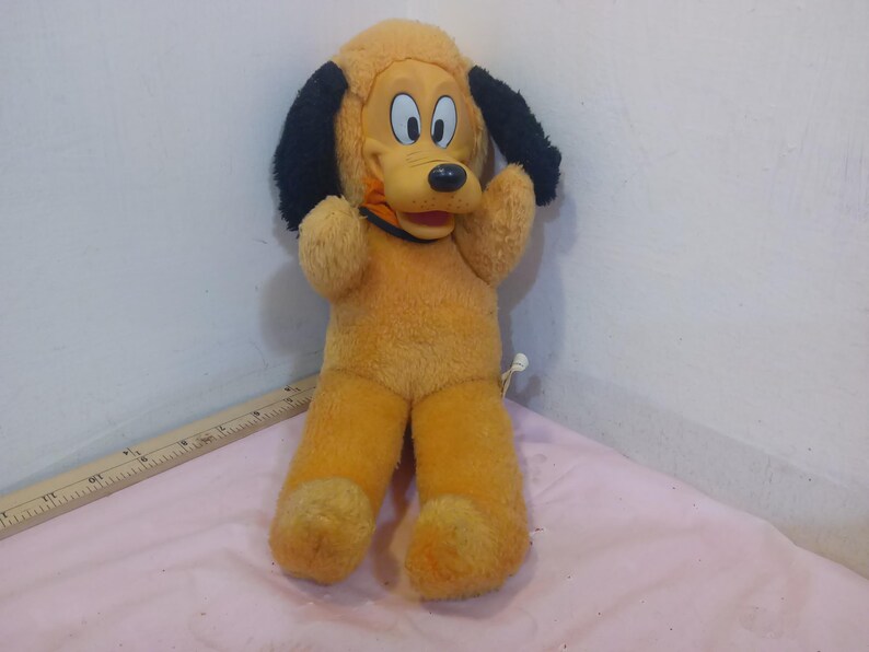 May include: A vintage plush toy of a cartoon dog with yellow fur, black ears, and a black nose. The dog has a red collar and is in a standing position with its arms raised. The toy is in good condition.