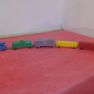 Puede incluir: Un tren de juguete con una locomotora azul, un vagón verde, un vagón gris, un vagón amarillo y un vagón rojo.