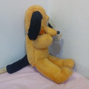 May include: A vintage Pluto plush toy, featuring a yellow body, black ears, and a black tail. The plush dog is in a seated position, with a smiling expression. The toy is made of soft, plush material.