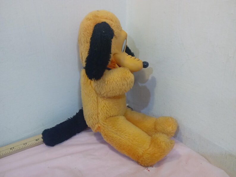 May include: A vintage Pluto plush toy, featuring a yellow body, black ears, and a black tail. The plush dog is in a seated position, with a smiling expression. The toy is made of soft, plush material.