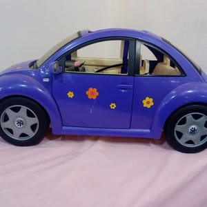 Puede incluir: Un coche de juguete morado con forma de Volkswagen Beetle con interior negro y ruedas plateadas. El coche tiene calcomanías de flores amarillas en el lateral.