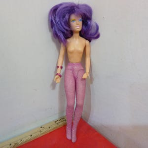 May include: A vintage doll with purple hair, blue eyes, and a nude torso. The doll is wearing pink, glittery leggings and matching bracelets. The doll is standing on a red surface.