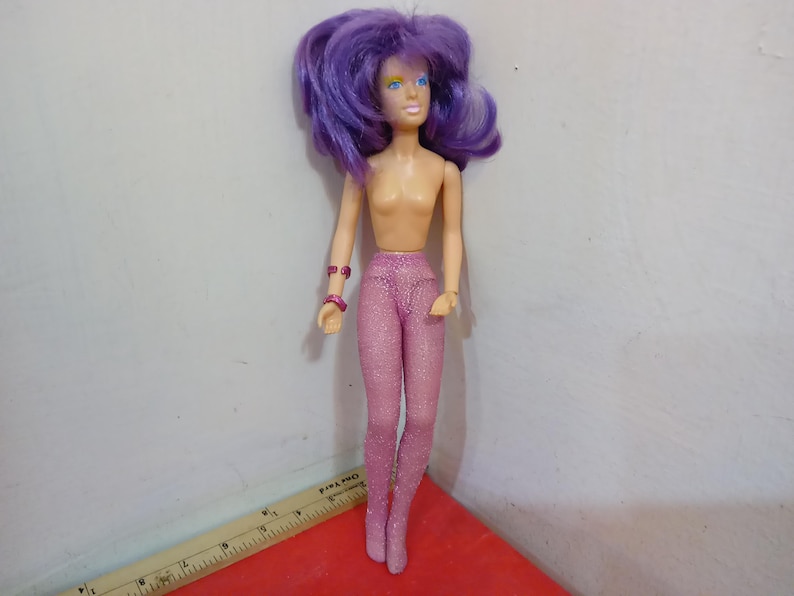 May include: A vintage doll with purple hair, blue eyes, and a nude torso. The doll is wearing pink, glittery leggings and matching bracelets. The doll is standing on a red surface.