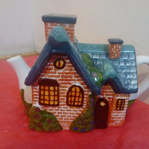 May include: A ceramic teapot shaped like a brick house with a blue roof, white spout, and brown door. The house has two windows with yellow glass and green vines growing on the side.