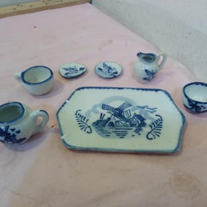 Puede incluir: Un juego de té de cerámica en miniatura con un diseño de molino de viento azul y blanco. El juego incluye una tetera, una crema, tazas, platillos y una bandeja. Las piezas están dispuestas sobre una superficie rosa.