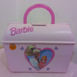 以下が含まれることがあります： ピンクのプラスチック製のバービーのドクターキット。ハンドルと、バービーが赤ちゃん人形を抱いているハート型の画像があります。キットには、前面に「バービー」という文字が印刷されています。