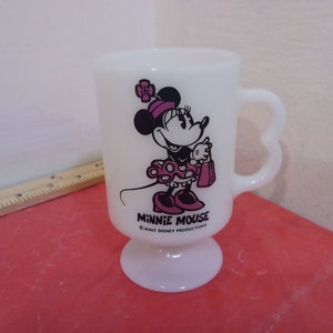 May include: White ceramic mug with a heart-shaped handle. The mug features a black and white illustration of Minnie Mouse wearing a pink dress and holding a pink shopping bag. The text "Minnie Mouse" and "&copy; WALT DISNEY PRODUCTIONS" are printed on the mug.