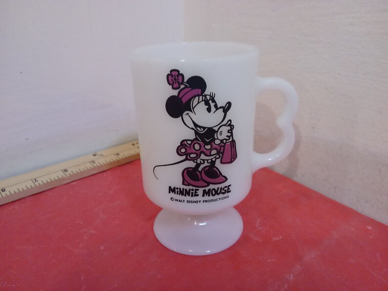 May include: White ceramic mug with a heart-shaped handle. The mug features a black and white illustration of Minnie Mouse wearing a pink dress and holding a pink shopping bag. The text "Minnie Mouse" and "&copy; WALT DISNEY PRODUCTIONS" are printed on the mug.