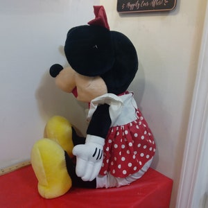 Puede incluir: Un gran juguete de peluche de Minnie Mouse que lleva un vestido de lunares rojos y blancos con un delantal blanco. Tiene un lazo rojo en la cabeza y zapatos amarillos.