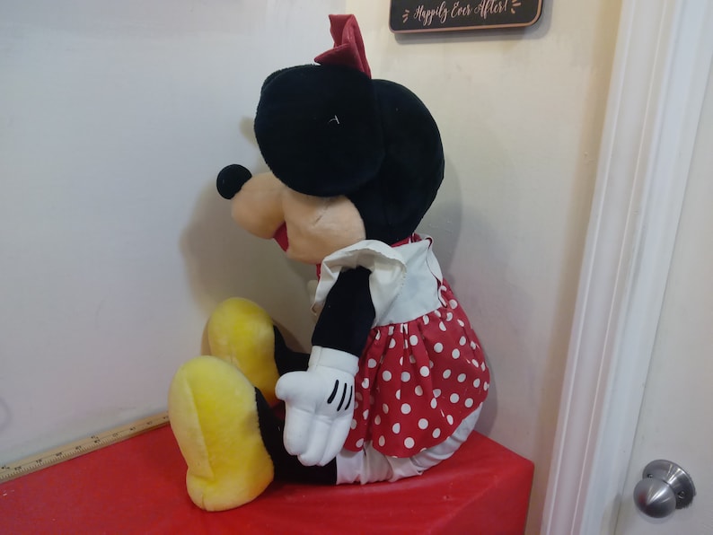 Puede incluir: Un gran juguete de peluche de Minnie Mouse que lleva un vestido de lunares rojos y blancos con un delantal blanco. Tiene un lazo rojo en la cabeza y zapatos amarillos.