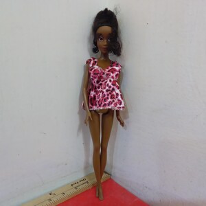 May include: A fashion doll with dark skin and black hair styled in a high bun. The doll wears a pink and white animal print top. The doll is standing on a red surface next to a ruler.