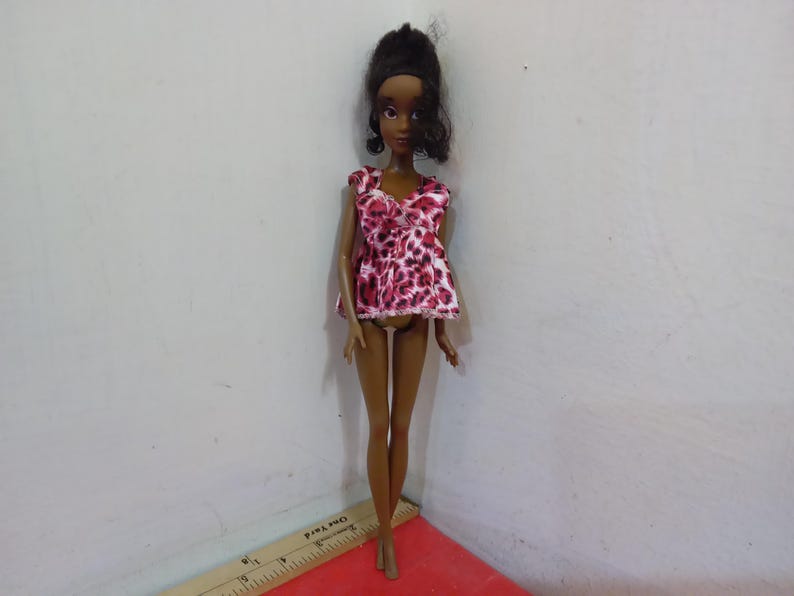 May include: A fashion doll with dark skin and black hair styled in a high bun. The doll wears a pink and white animal print top. The doll is standing on a red surface next to a ruler.