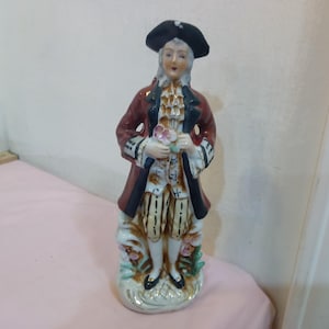 May include: A porcelain figurine of a man in a red and black coat, white shirt, and striped pants. He is holding a bouquet of flowers and wearing a black tricorne hat. The figurine is standing on a white base with floral details.