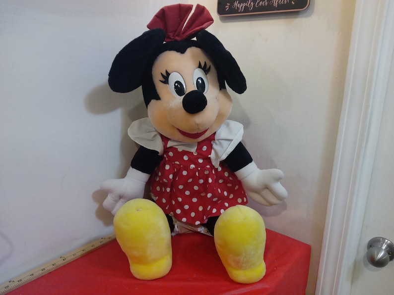Puede incluir: Un gran juguete de peluche de Minnie Mouse que lleva un vestido rojo con lunares blancos y un lazo rojo. El juguete tiene orejas negras, zapatos amarillos y un cuello blanco.