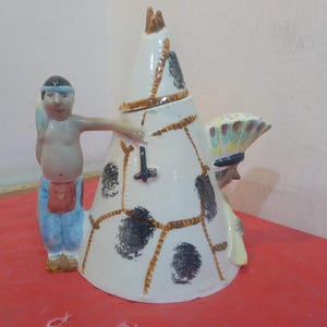May include: A ceramic teapot shaped like a conical teepee with a white base, brown painted structural lines, and black circular patterns. One side features a figure in blue trousers and a headband, while the other side shows a figure with a headdress.
