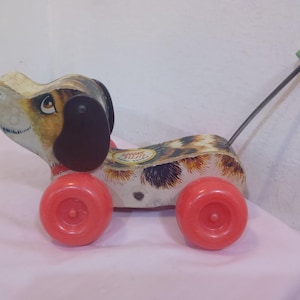 Op de afbeelding: Een vintage houten trekdier in de vorm van een hond met een gevlekte bruine en witte vacht. De hond heeft zwarte oren en een rode neus. Het speelgoed heeft rode wielen en een groene trekkoord.