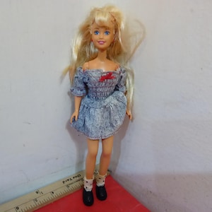 May include: A doll with blonde hair and blue eyes, wearing a denim-style dress with a red bow and black shoes. The doll has white socks with black polka dots. The doll is standing on a red surface.