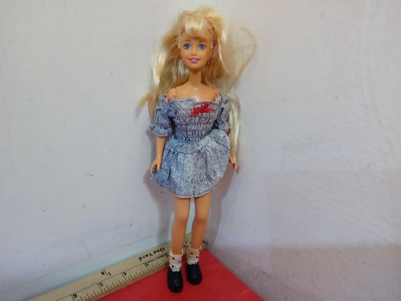 May include: A doll with blonde hair and blue eyes, wearing a denim-style dress with a red bow and black shoes. The doll has white socks with black polka dots. The doll is standing on a red surface.