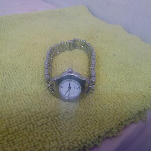 May include: A silver wristwatch with a white face and black hands and numerals. The watch has a linked metal strap and is set against a yellow-green textured surface. The watch face shows the time.