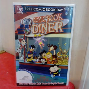 Op de afbeelding: Een stripboekcover met de titel "COMIC BOOK DINER" en de tekst "FREE COMIC BOOK DAY". De cover bevat cartoonfiguren en het websiteadres kidslovecomics.com.