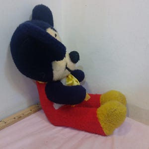 May include: A vintage Mickey Mouse plush toy, featuring a black head and ears, a white face, and a red body. The plush has yellow feet and a yellow bow tie. The toy is in a seated position.