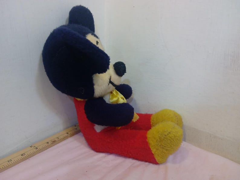May include: A vintage Mickey Mouse plush toy, featuring a black head and ears, a white face, and a red body. The plush has yellow feet and a yellow bow tie. The toy is in a seated position.