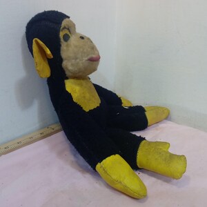 May include: A vintage stuffed monkey toy with black fur, yellow accents on the ears, hands, and feet, and a tan face. The monkey has a pink mouth and black eyes. The toy is lying on a pink surface.