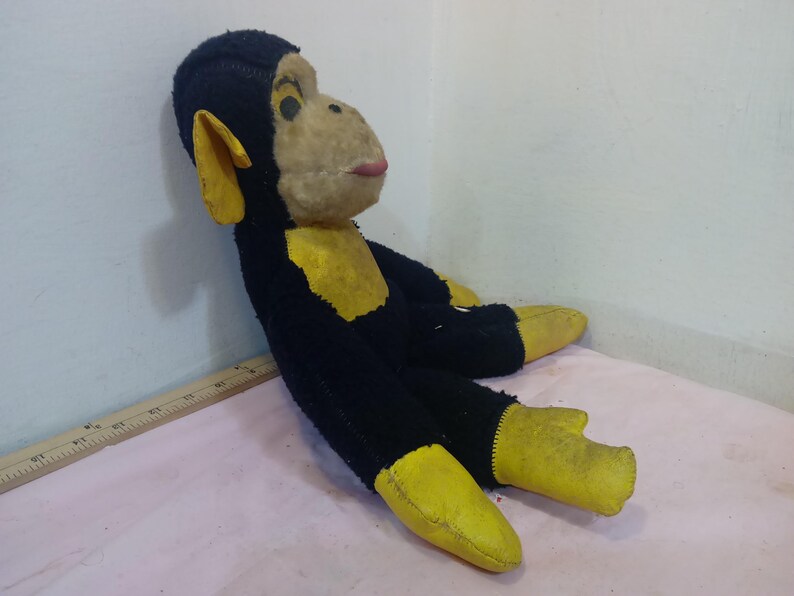 May include: A vintage stuffed monkey toy with black fur, yellow accents on the ears, hands, and feet, and a tan face. The monkey has a pink mouth and black eyes. The toy is lying on a pink surface.