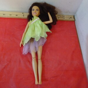 May include: A doll with brown hair and a light complexion wearing a light green and lavender dress. The doll is posed on a red surface. The doll's arms are raised, and the legs are straight.