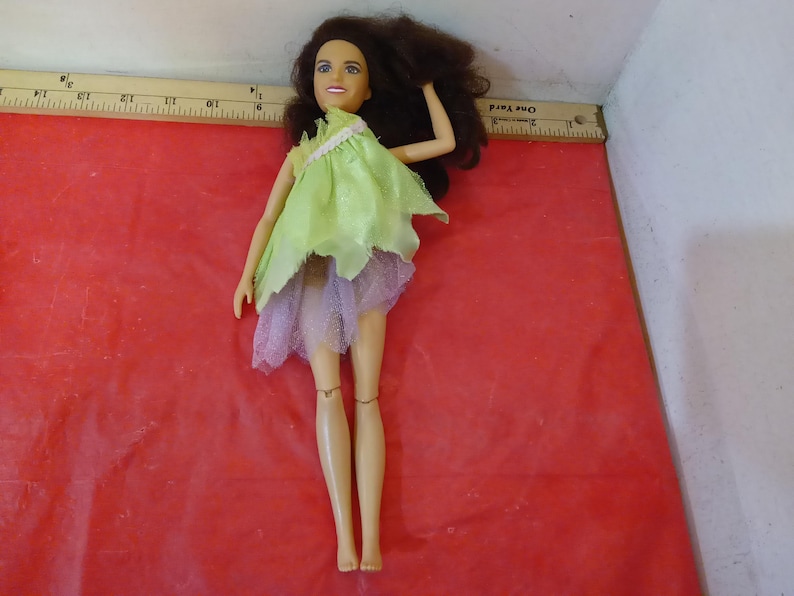 May include: A doll with brown hair and a light complexion wearing a light green and lavender dress. The doll is posed on a red surface. The doll's arms are raised, and the legs are straight.
