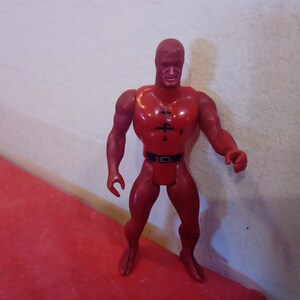 Pode incluir: Figura de a&ccedil;&atilde;o de pl&aacute;stico vermelho de um super-her&oacute;i vestindo um traje vermelho com um cinto preto e uma m&aacute;scara preta. A figura tem uma constru&ccedil;&atilde;o muscular e est&aacute; de p&eacute; com os bra&ccedil;os ao lado do corpo.