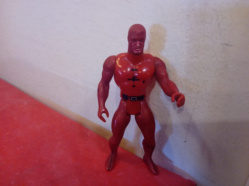 Pode incluir: Figura de a&ccedil;&atilde;o de pl&aacute;stico vermelho de um super-her&oacute;i vestindo um traje vermelho com um cinto preto e uma m&aacute;scara preta. A figura tem uma constru&ccedil;&atilde;o muscular e est&aacute; de p&eacute; com os bra&ccedil;os ao lado do corpo.