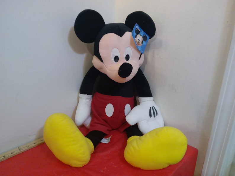 May include: A large plush Mickey Mouse toy sitting on a red surface. Mickey is wearing his signature red shorts with two white buttons and yellow shoes. He has black ears and a black nose.