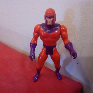 Pode incluir: Uma figura de a&ccedil;&atilde;o vermelha e roxa de um super-her&oacute;i com capacete. A figura tem uma constru&ccedil;&atilde;o muscular e est&aacute; de p&eacute; com os bra&ccedil;os ao lado do corpo.
