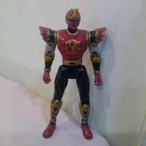 Puede incluir: Una figura de acción roja y negra de un Power Ranger con detalles dorados. La figura tiene un casco con una visera roja y una pieza de pecho roja con un símbolo dorado. La figura está de pie sobre una superficie blanca.