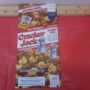 Könnte beinhalten: Eine Vintage-Cracker-Jack-Box mit karamellbeschichtetem Popcorn und Erdnüssen. Die Box hat ein rot-weißes Design mit dem Schriftzug "Cracker Jack". Sie zeigt einen Seemann und seinen Hund. Nettogewicht: 28,3 g.