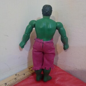 May include: Green plastic action figure of the Hulk wearing a pink and purple loincloth. The figure is posed with its arms at its sides and its fists clenched.