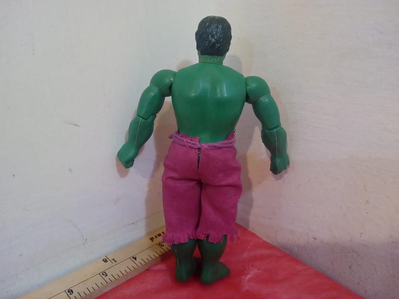 May include: Green plastic action figure of the Hulk wearing a pink and purple loincloth. The figure is posed with its arms at its sides and its fists clenched.