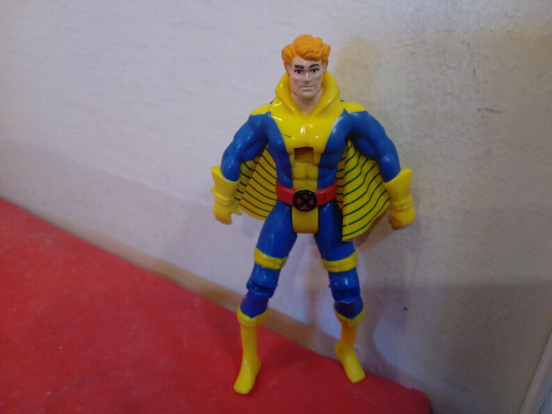 Pode incluir: Uma figura de a&ccedil;&atilde;o azul e amarela com uma capa amarela e um cinto vermelho. A figura tem uma cabe&ccedil;a amarela com cabelo laranja e uma express&atilde;o s&eacute;ria.