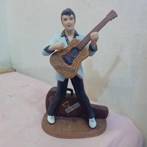 May include: A figurine of a man playing a guitar. The figure is wearing a light blue shirt, black trousers, and white shoes. The guitar is brown with decorative detailing and the words "ELVIS PRESLEY" on it. A brown suitcase with the word "MEMPHIS" is behind the figure.