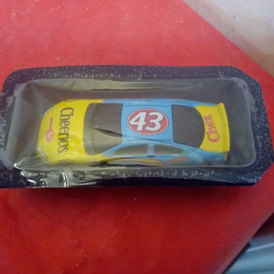 May include: A blue and yellow toy race car with the number 43 and the word "Cheerios" on the side. The car is in a plastic package.