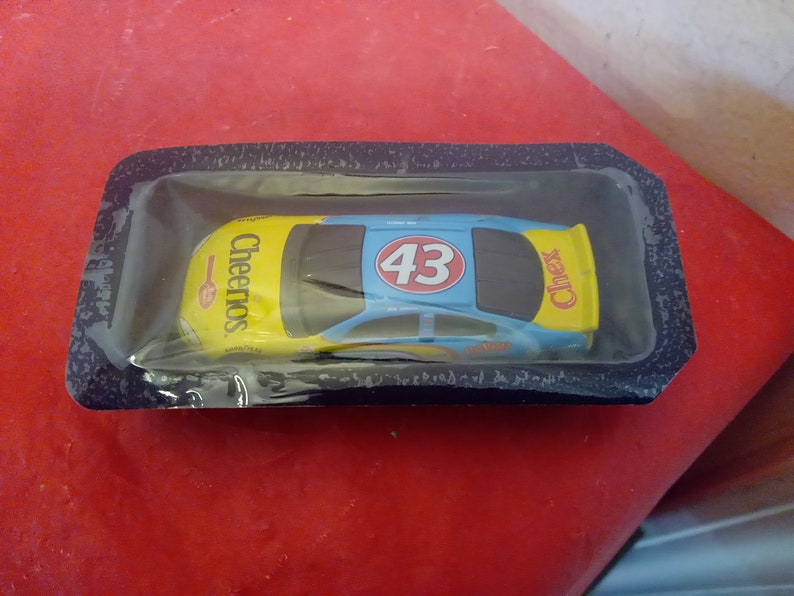 May include: A blue and yellow toy race car with the number 43 and the word "Cheerios" on the side. The car is in a plastic package.