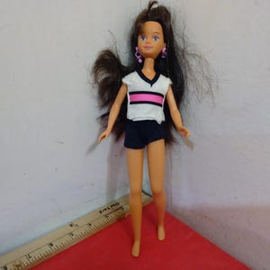 May include: A doll with long brown hair, wearing a white and black top with a pink stripe, and dark blue shorts. The doll has pink earrings and is standing on a red surface. A ruler is in the foreground.