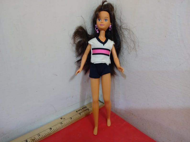 May include: A doll with long brown hair, wearing a white and black top with a pink stripe, and dark blue shorts. The doll has pink earrings and is standing on a red surface. A ruler is in the foreground.