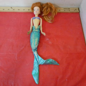 May include: A mermaid doll with auburn hair and a blue and purple top. The doll has a turquoise tail with a shimmering effect. The doll is lying on a red surface.