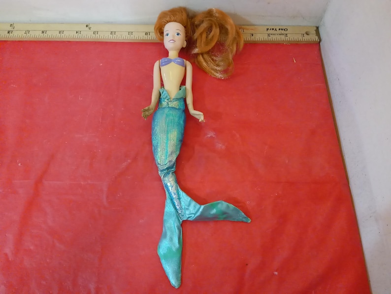 May include: A mermaid doll with auburn hair and a blue and purple top. The doll has a turquoise tail with a shimmering effect. The doll is lying on a red surface.