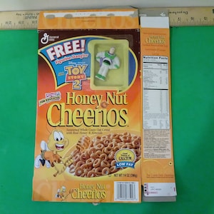 Puede incluir: Una caja de cereal Honey Nut Cheerios con una figura de Buzz Lightyear de Toy Story 2 gratis. La caja es amarilla y naranja con una imagen de una abeja en la parte delantera. El cereal es bajo en grasa y una buena fuente de calcio. La caja contiene 396 gramos.