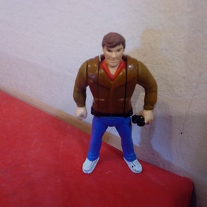 Pode incluir: Uma jaqueta de couro marrom com uma camisa vermelha por baixo, jeans azul e t&ecirc;nis branco. A figura est&aacute; segurando um objeto preto na m&atilde;o direita.